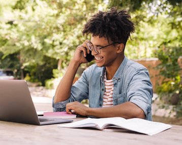 A young man with curly hair is sitting at an outdoor table, talking on the phone and smiling. In front of him are a laptop and some books.