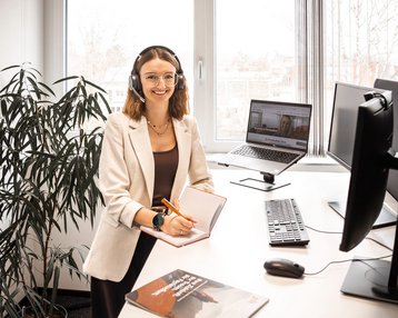 A woman with a headset sits at a desk, working on a computer, surrounded by plants in a modern office.