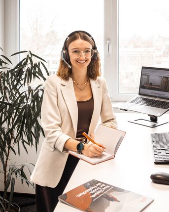 A woman with a headset sits at a desk, working on a computer, surrounded by plants in a modern office.