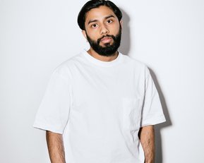 A man with a beard is wearing a white T-shirt and black pants. He stands against a neutral background, with his hands casually in his pockets.