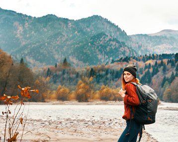 A woman with a backpack stands by the riverbank, surrounded by autumn trees and mountains. She is wearing a red sweater and a beanie, smiling at the camera.