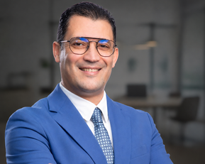 A man in a blue suit with glasses smiles as he stands with his arms crossed in front of a modern office backdrop.