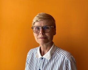 A woman with short blonde hair and a striped blouse stands in front of an orange background. She is wearing stylish glasses.