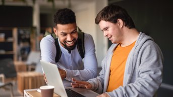 Two men are standing at a table, looking at a laptop together. One of the men is smiling while the other points at the screen. There’s a coffee cup on the table.
