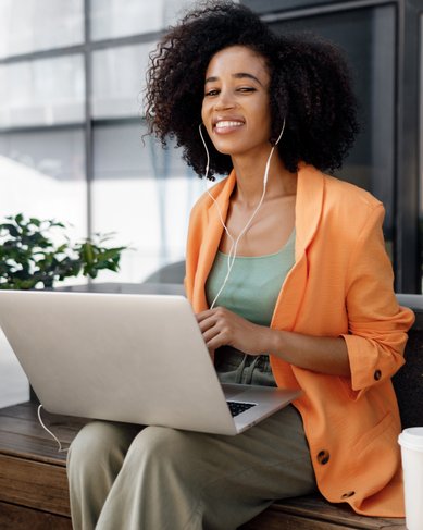 A woman with curly hair is sitting at an outdoor table, wearing an orange blazer and listening to music while working on her laptop.