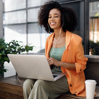 A woman with curly hair is sitting at an outdoor table, wearing an orange blazer and listening to music while working on her laptop.