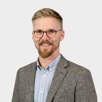 A man with blonde hair and glasses is wearing a gray blazer over a striped shirt. He is smiling warmly and standing against a neutral background.