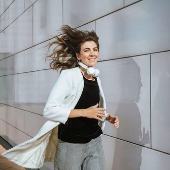 A woman with headphones walks along a wall, wearing a white blazer and has long, flowing hair.