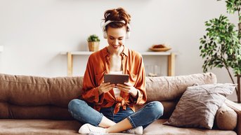 A woman is sitting on a sofa, wearing headphones and smiling as she holds a tablet in her hands. She is dressed in an orange top and jeans. In the background, plants and a shelf are visible.