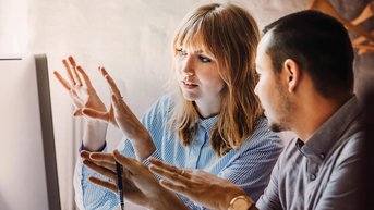 A woman and a man are discussing at a computer while gesturing. The woman is wearing a striped shirt and the man is in a gray polo shirt. They both seem to be working on a creative project.