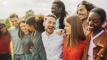 A group of nine people stands closely together, smiling. They are wearing various casual outfits and seem to be having a great time together. The scene conveys a sense of community and friendship.