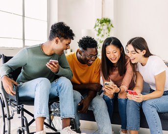 Four young people are sitting on a sofa, looking at their smartphones together. One of the participants is in a wheelchair. They all seem engaged and are laughing with each other.