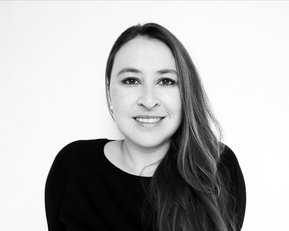 A woman with long brown hair and a friendly smile is wearing a black top. The background is neutral and bright.