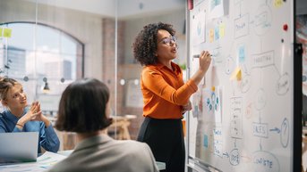 A woman with curly hair is presenting at a whiteboard while two other people listen attentively. The whiteboard displays diagrams and notes that represent various ideas and concepts.